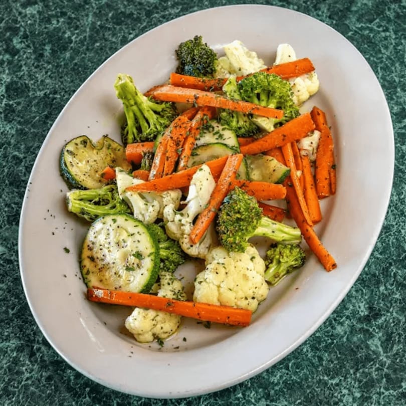 A white oval plate filled with roasted broccoli, cauliflower, carrots, and zucchini on a green marble surface.