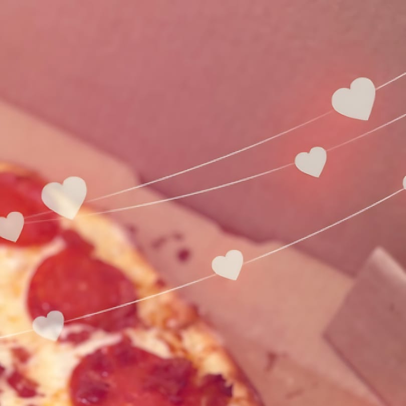 Pepperoni pizza in a box with heart-shaped decorations on strings, suggesting a romantic theme.