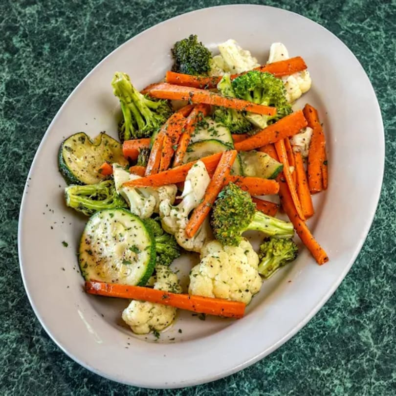 A white oval plate filled with steamed carrots, zucchini, broccoli, and cauliflower on a green marble surface.