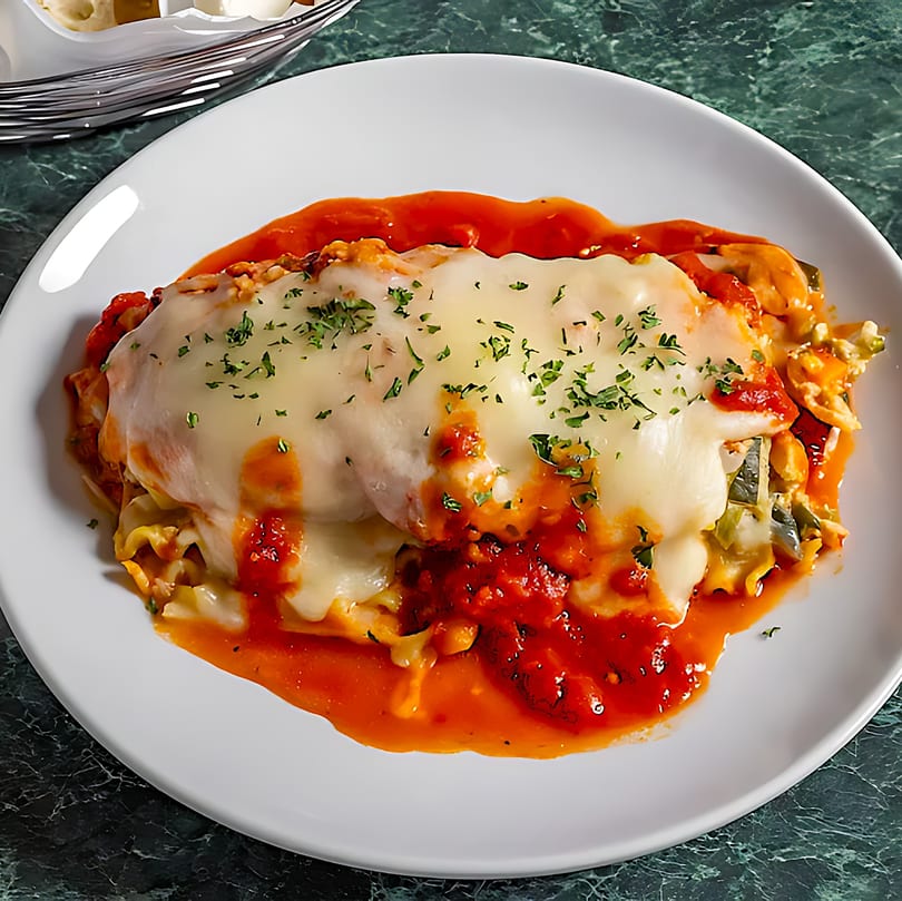 A white plate holds a serving of lasagna topped with melted cheese, red sauce, and parsley.